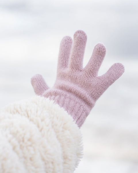 A hand wearing a light pink knitted glove with a ribbed wrist extends upwards and slightly to the left, revealing a plush, cream-colored sleeve on the arm, against a blurred light background.