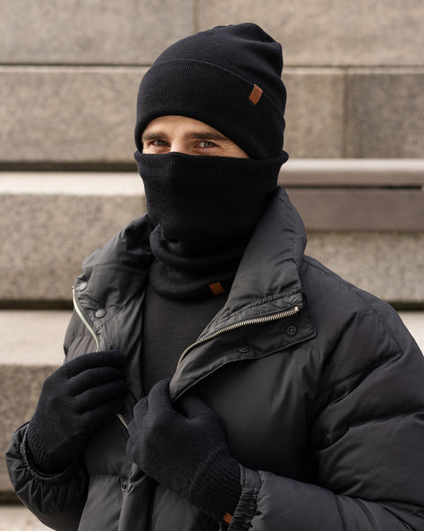 A person, who appears to be male, is standing outdoors on concrete steps, wearing a black knit beanie, a black knit neck gaiter pulled up over their mouth and nose, black knit gloves, and a black puffer jacket. Only their eyes are visible above the neck gaiter. The beanie has a small brown tag on the folded brim. The person seems to be looking directly at the camera.