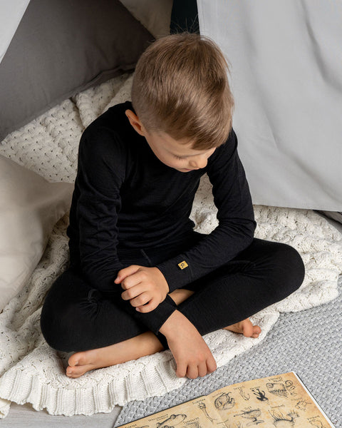 A boy wearing the menique Kids' Long Sleeve Set 160 Merino in Black color, reading a book, sitting on a carpet. 