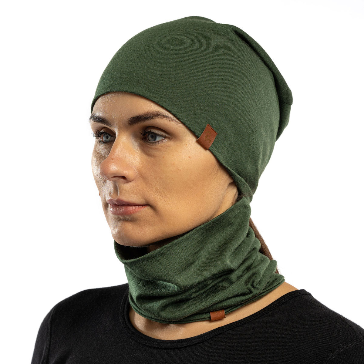 The picture features a woman wearing the unisex gaiter and beanie merino set in dark green color. She has her hands raised above her head. She is in a forest.